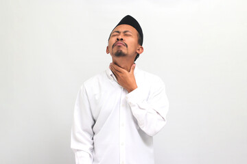 Sad Young Indonesian man, wearing a songkok, peci, or kopiah, holding his neck, suffering from a sore throat, cold, or flu, isolated on white background