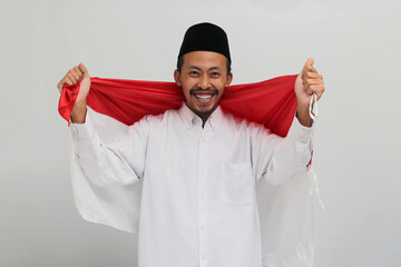 an Excited young Indonesian man, wearing a songkok, peci, or kopiah, is holding an Indonesian flag...