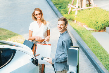 Young couple travel with EV electric car charging in green sustainable city outdoor garden in...