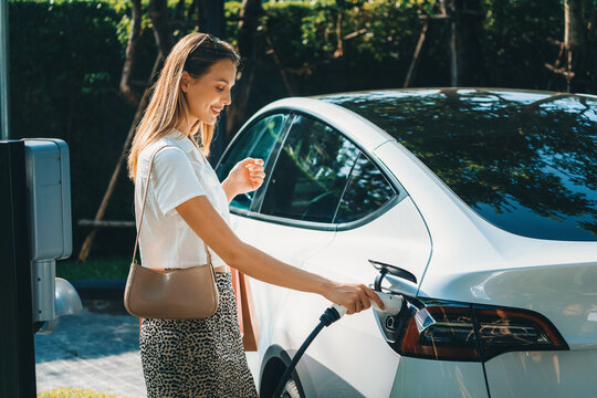 Young woman travel with EV electric car charging in green sustainable city outdoor garden in summer. Urban sustainability lifestyle by green clean rechargeable energy of electric BEV vehicle innards