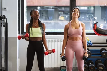 Küchenrückwand Plexiglas Fitness two fitness women working out with weights in the gym and looking at the camera with a smile on their faces