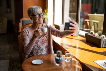 Mature woman is drinking coffee in cafe. She is taking selfie.