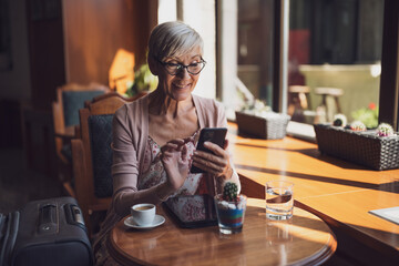 Mature woman is sitting in a cafe at the airport, waiting for boarding. She is examining schedule on smartphone.