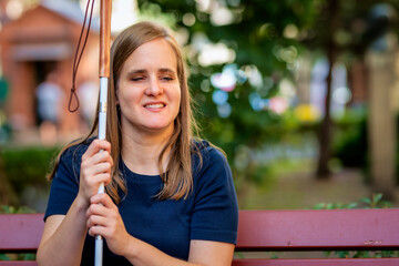 A visually impaired woman holding a white cane and sitting on a bench in the city - 649166337