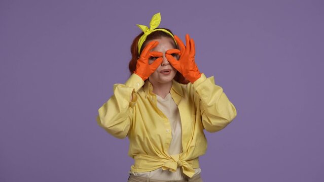 Woman in casual clothing and rubber gloves jokingly looking through fingers as binocles, smiling at the camera. Isolated on purple background.