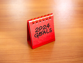 Start a new year 2024 banner background. Handwritten text, goals and 2024 numbers year with target icon on red small desk calendar cover standing on wooden table. Business goals and success concepts.