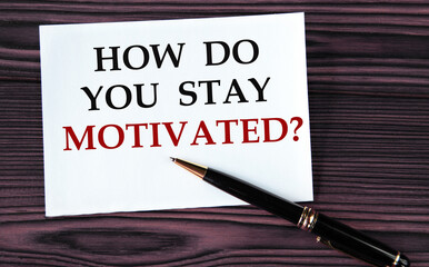 HOW DO YOU STAY MOTIVATED? - words on a white sheet on a brown wooden background with a pen