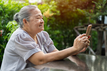 Fototapeta premium Happy old grandmother holding mobile phone, video call with loved ones while relaxing in the garden at home.