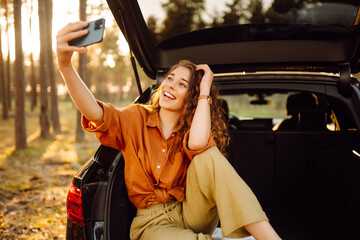 A young woman admires the sunset in the trunk of a car with a phone in her hands. A curly-haired tourist takes pictures on a mobile phone and communicates via video call. Travel, weekend concept.