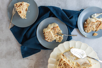 Flat lay or top shot of three tier hummingbird or carrot cake with cream cheese frosting and pecan nuts or walnuts on a white cake stand with slices being served on plates