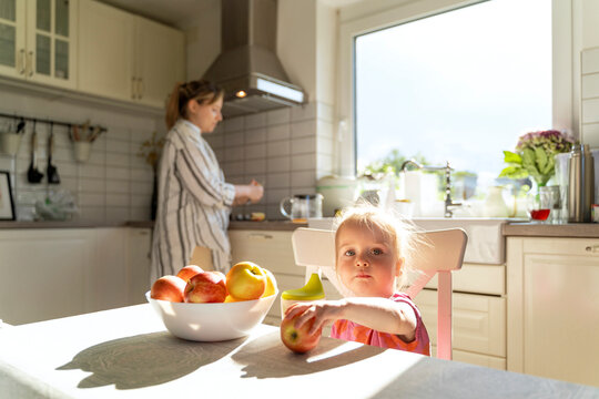 Girl holding apple fruit at dining table with mother cooking in kitchen