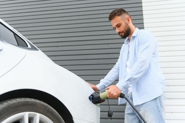 Beautiful young stylish man is with electric car at daytime charging the vehicle