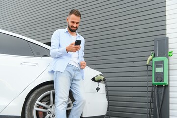 Happy man using smart phone and charging car at vehicle charging station