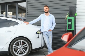 Beautiful young stylish man is with electric car at daytime charging the vehicle