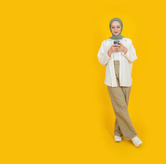 Full body young caucasian muslim girl in abaya hijab holding using modern mobile cell phone smartphone isolated on yellow studio background spy space. Islam religious lifestyle concept idea image.