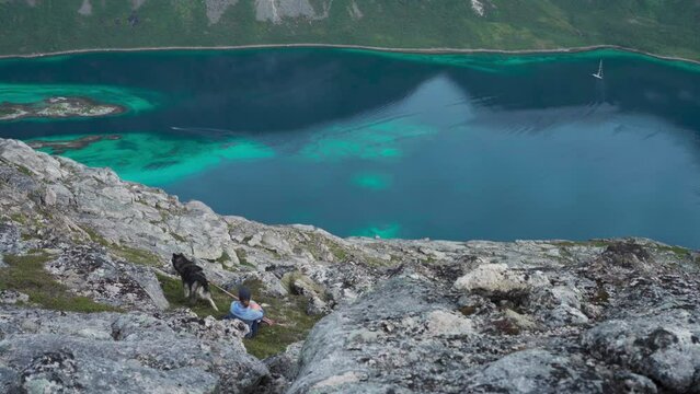 Female Wanderlust Sitting On Rocky Hill Salberget With Pet Dog Overlooking Calm Blue Lake In Norway. high angle
