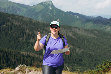 Stylish sport woman holding paper map, wearing backpack and hat looking at mountain view while...