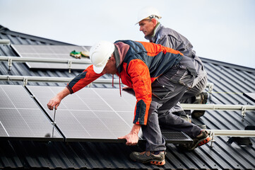 Builders installing photovoltaic solar panels on roof of house. Men engineers in helmets building solar module system with help of hex key. Concept of alternative, renewable energy. - obrazy, fototapety, plakaty