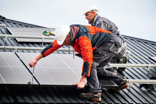 Builders installing photovoltaic solar panels on roof of house. Men engineers in helmets building solar module system with help of hex key. Concept of alternative, renewable energy.