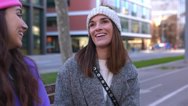 Two female friends in wooly hat hanging out sitting on bench in city street. Young millennial women talking while taking a break outdoors in winter season. Friendship and people concept.