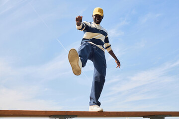 Minimal fashion shot of young black man wearing trendy outfit standing on roof against blue sky and...