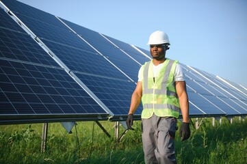 African american man in white helmet and grey overalls standing among rows of solar panels