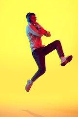 Fototapeta na wymiar Full lenght portrait of thinking young guy listening music in headphones and jumping against on yellow background in red color light, filter.