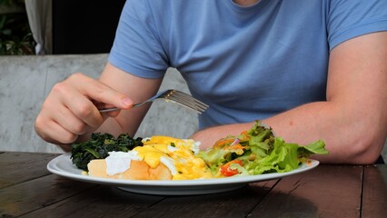Man eating healthy food as poached egg, salad, toast in cafe. Close-up of man pierces tomato with fork dips in yolk on poached egg. Order poached egg in cafe to eat healthy food. Concept healthy food.