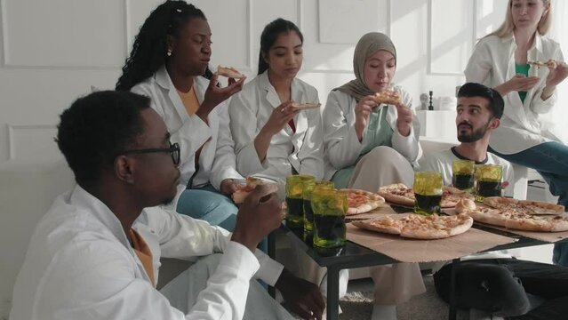 Young diverse multiethnic students interns doctors having a break from work, sitting at office, chatting and eating pizza meal lunch. Colleagues spending time together during lunch time. Pizza time 