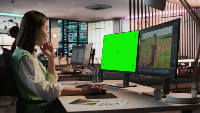 Female Asian Game Designer Using Desktop Computer With Green Screen Chromakey on Display, Designing Characters In 3D Modeling Software For Survival Video Game. Woman Working In Game Development Office