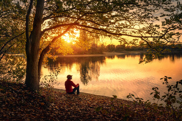 Man sitting under a tree and enjoying a tranquil beautiful sunset at a lake alone, with warm glowing colors - 649209537