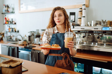 Smiling woman barista works in a cafe. A young woman in an apron behind the bar holds to-go coffee and desserts for ordering.
