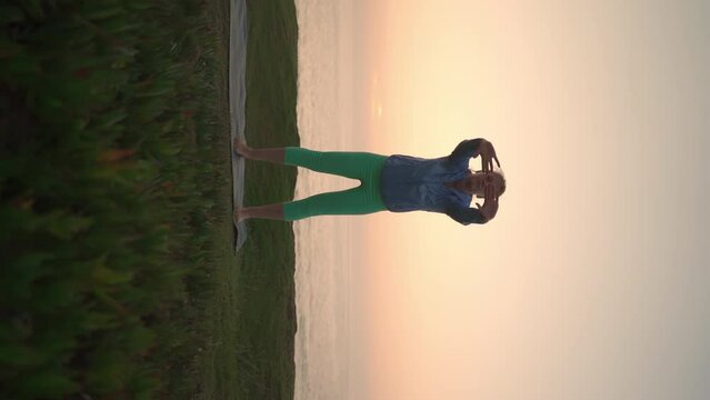 Senior woman doing qigong exercises outdoors. Sports activities on fresh air at sunset. Pensioner woman practicing qigong barefoot on grass on ocean shore. Body care, wellness, physical activity