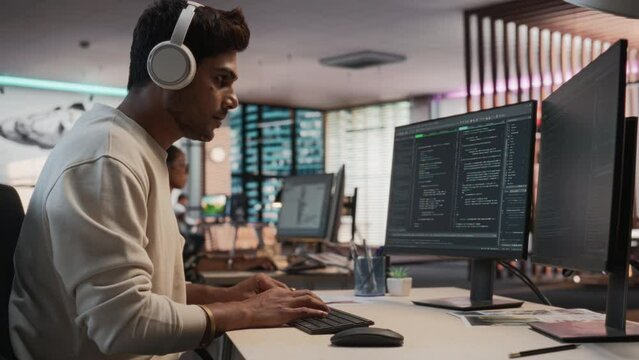 Male Indian Game Programmer Coding On Desktop Computer In Game Development Studio Diverse Office. Focused Man Writes Lines Of Code, Does Gameplay Engineering For New Immersive 3D RPG Video Game.