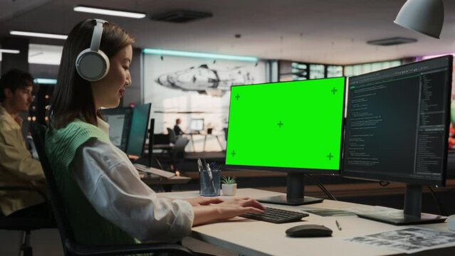 Female Asian Game Programmer Coding On Desktop Computer With Green Screen Chromakey On Display In Game Design Studio Office. Stylish Woman Does Gameplay Engineering For New Immersive 3D RPG Video Game