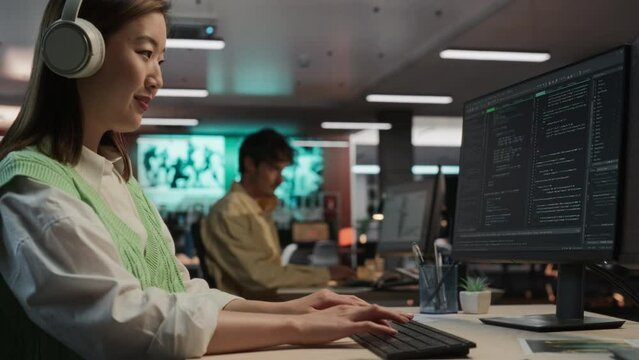 Female Asian Game Programmer Coding On Desktop Computer In Game Design Studio Diverse Office. Woman Listening to Music, Writes Lines Of Code. Gameplay Engineer of New Immersive 3D Survival Video Game.