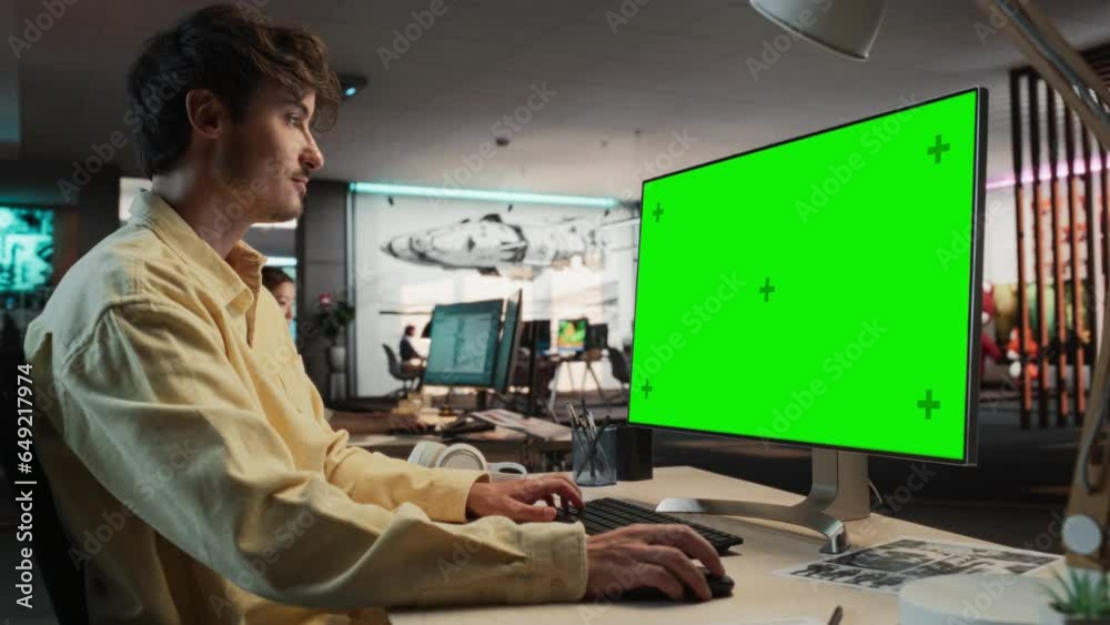 Poster Portrait of Caucasian Man Sitting at His Desk Using Desktop Computer with Mock-up Green Screen Chromakey. Male Gameplay Animator Working in Game Design Startup, Finishing Details on a Project.