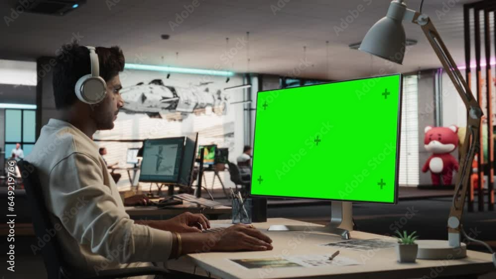 Poster Portrait of Indian Man Sitting at His Desk Using Desktop Computer with Mock-up Green Screen Chromakey. Male Gameplay Animator Working in Game Design Startup Office, Finishing Details on a Project.