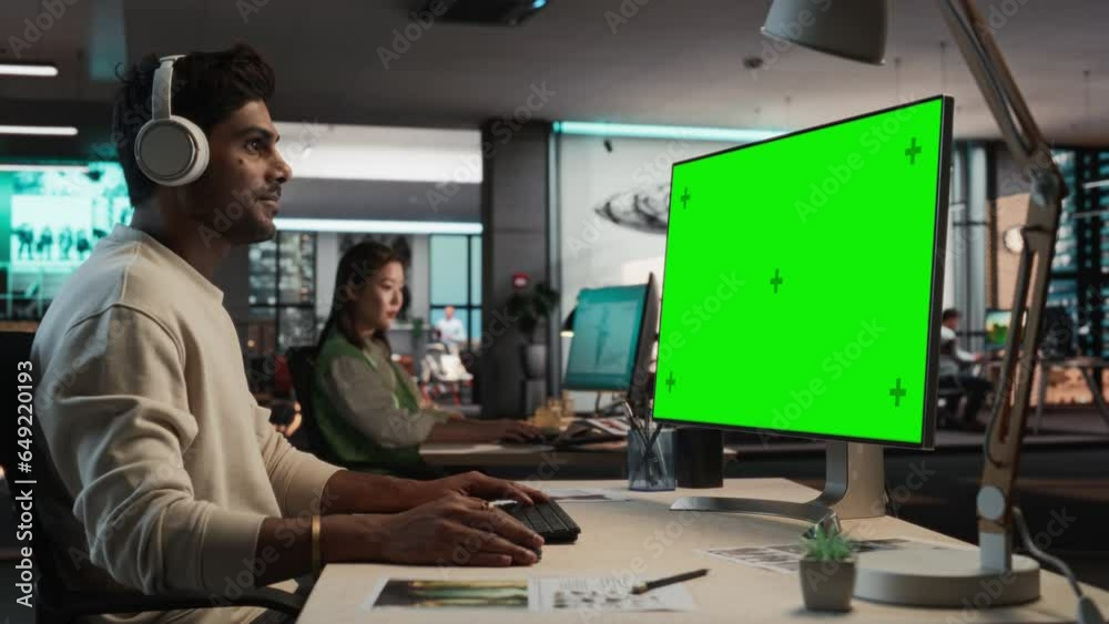 Poster Portrait of Creative Indian Man Sitting at a Desk Using Desktop Computer with Mock-up Green Screen Chromakey. Male Concept Artist Working in Game Design Company Office, Creating Immersive Gameplay.