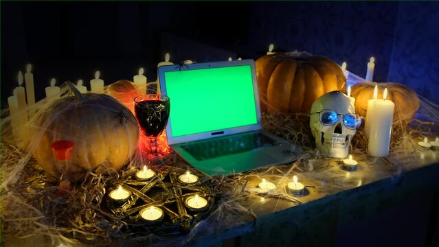Altar Halloween party attributes, laptop with chromakey on screen traditional pumpkins flickering candles glass of red wine old skull next to pentagram among hay in barn. Mystical horror background.