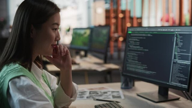 Female Asian Game Programmer Coding On Desktop Computer In Game Development Studio Diverse Office. Focused Woman Writes Lines Of Code, Does AI Engineering For Playable Characters 3D RPG Video Game.