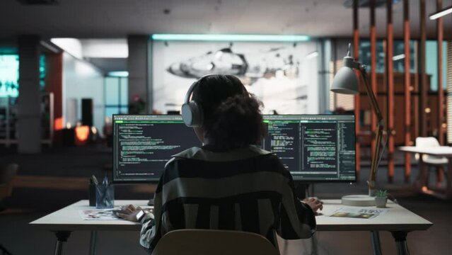 Creative Office: Professional Black Female Programmer Uses Headphones, Working on Desktop Computer. Focused Coder, Software Engineer Developing App, Video Game. Listening to Podcast, Music. Back View.