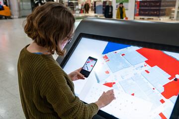 Woman with phone uses self-service kiosk in the shopping mall. - obrazy, fototapety, plakaty