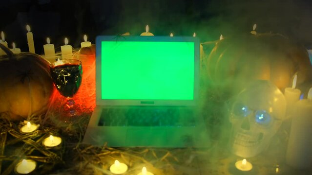 Halloween time, in smoke crypt vampire, glass of blood and laptop with chromakey green screen, pumpkins, flickering candles, human skull in cobweb, spirits night. Eerie scary spooky fall decoration.