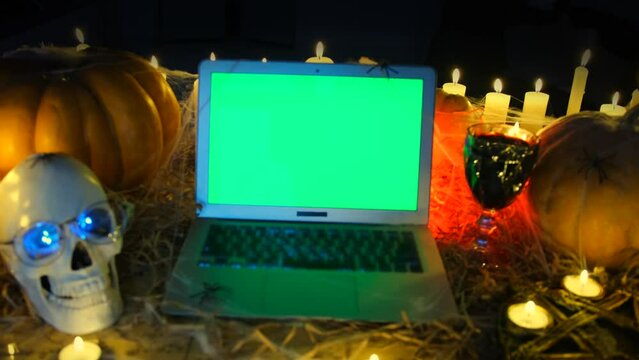 Zooms in shot, creepy vampire crypt laptop with chromakey screen pumpkins, candles, spooky skull, in darkness, mysterious holiday background in yellow-red light, close up. Halloween party invitation.