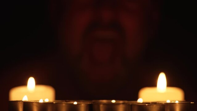 Burning candles with terrible man looks at camera and laughs terribly on background. Mentally ill scary male grimaces looking into camera. Spooky Halloween celebration october festival