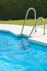 Swimming pool curb with ladder, with cool blue looking water and a bush fence in the background. There is green grass, in the summer.