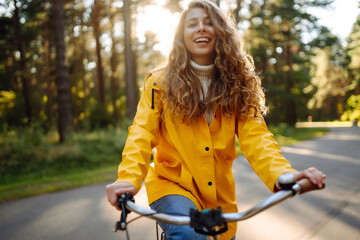 A smiling female tourist in a yellow coat enjoys the weather in the autumn park while riding a bicycle. Autumn fashion. Concept of relaxation, nature.