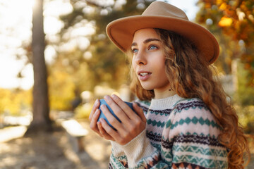 A happy woman in a stylish sweater sits on a bench in an autumn park, drinks a hot drink from a thermos and enjoys the view of the lake. Vacation concept, weekend. Lifestyle.