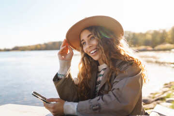 Cheerful woman in a hat relaxes, drinks tea or coffee under the sunlight against the backdrop of a lake. The concept of relaxation, freedom.